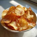 A close-up of a bowl filled with golden, crispy homemade potato chips sprinkled with salt.