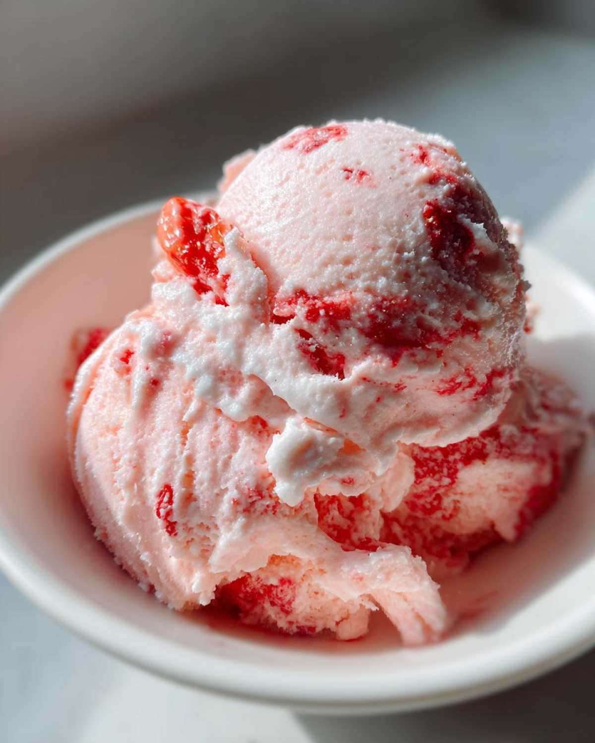 Close-up of two scoops of pink, homemade strawberry ice cream with visible red fruit pieces served in a white bowl.