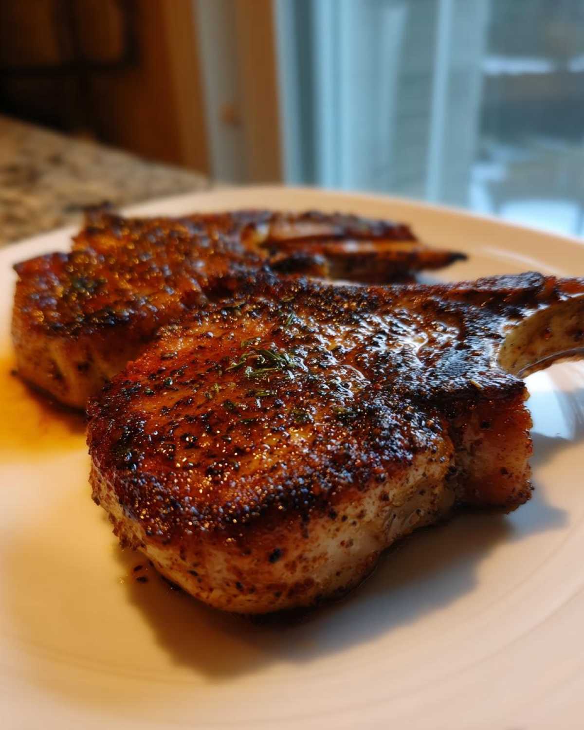 Close-up of two juicy baked pork chops with a dark, seasoned crust resting on a white plate.