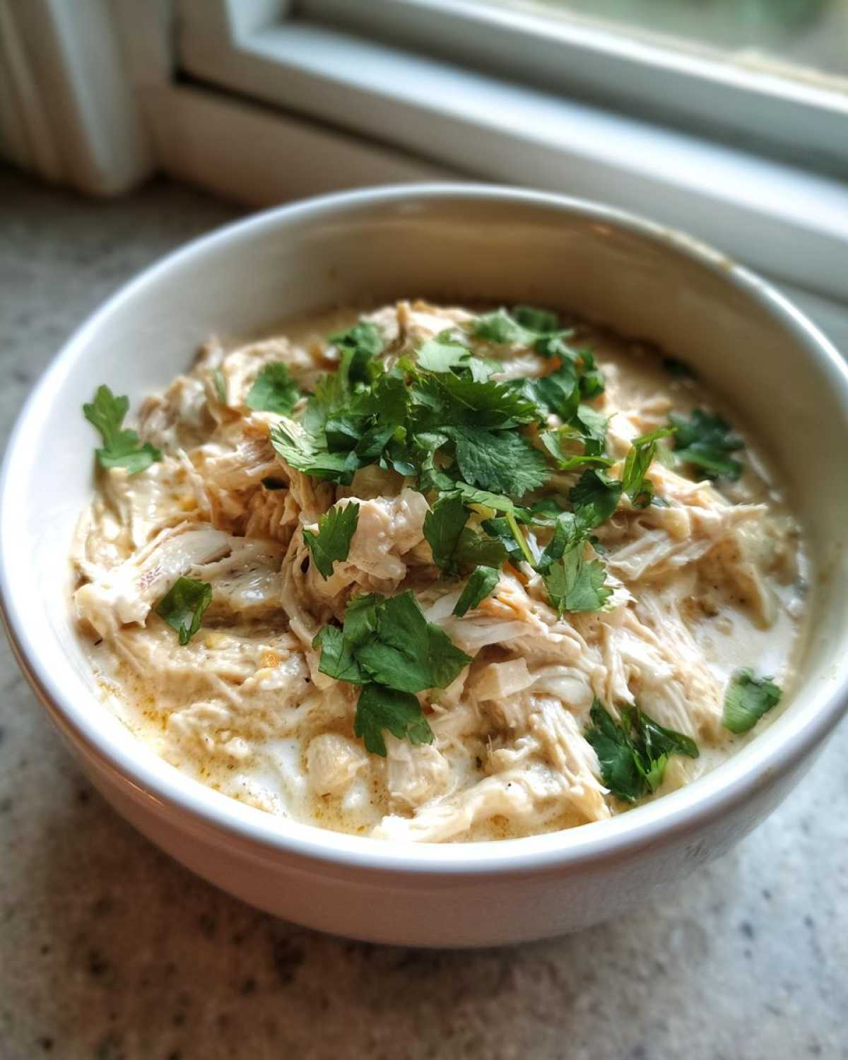 A close-up of a white bowl filled with creamy keto white chicken chili, featuring shredded chicken and fresh cilantro garnish.