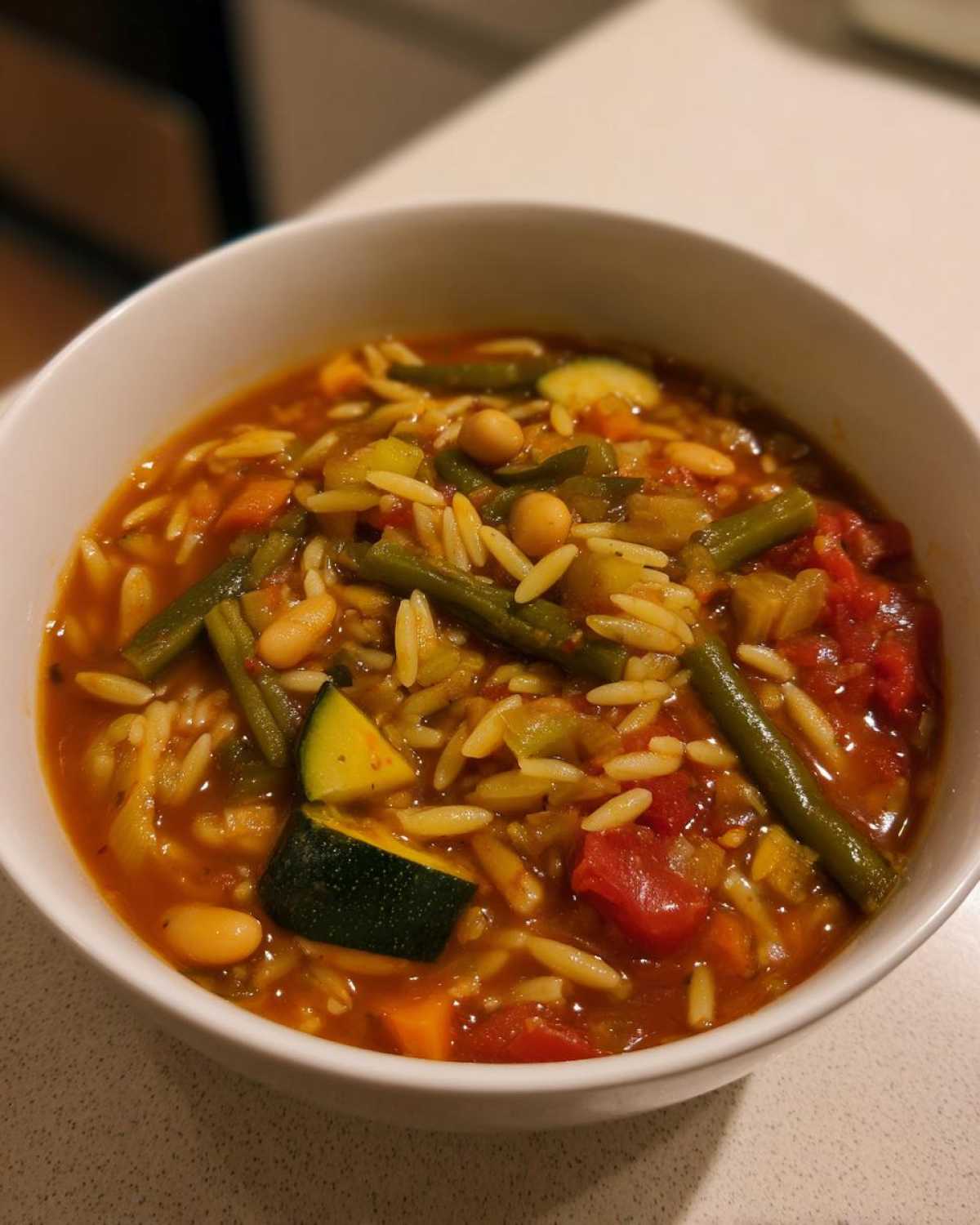 Close-up of a white bowl filled with vegetable minestrone soup featuring orzo pasta, zucchini, green beans, and tomatoes.