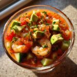 Close-up overhead view of a glass filled with vibrant mexican shrimp cocktail, featuring shrimp, avocado, and tomato sauce.