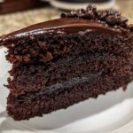 Close-up of a moist slice of chocolate fudge cake layered with dark ganache and topped with chocolate chips.