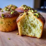 Close-up of a bitten pistachio muffin showing the moist interior and green pistachio pieces, topped with sugar and chopped pistachios.