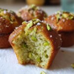 A close-up photo of a pistachio muffin broken in half, showing the moist crumb and whole pistachios inside. Other pistachio muffins are blurred in the background.