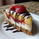 A decadent slice of no-bake banana split cake featuring a graham cracker crust, creamy filling, bananas, strawberries, and chocolate drizzle.
