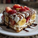 A decadent slice of no-bake banana split cake layered with cream, strawberries, and bananas, drizzled with chocolate sauce.