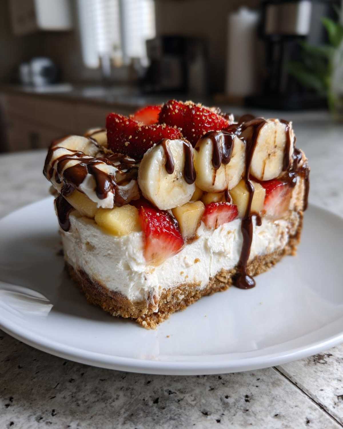 A thick slice of no-bake banana split cake featuring a graham cracker crust, creamy filling, and topped with bananas, strawberries, and chocolate drizzle.