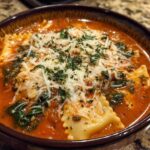 A close-up of a rich tomato broth one pot lasagna soup filled with ravioli, spinach, and topped with shredded cheese and parsley.
