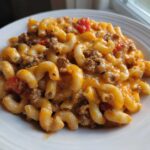 Close-up of a serving of cheesy taco pasta with elbow macaroni, ground beef, and tomatoes on a white plate.