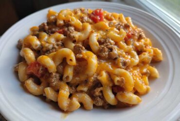 Close-up of a serving of cheesy taco pasta with elbow macaroni, ground beef, and tomatoes on a white plate.
