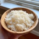Close-up of perfectly cooked, fluffy sushi rice served in a small wooden bowl held by a hand.
