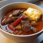 Close-up of a white bowl filled with rich, reddish-brown philly cheese steak soup featuring chunks of beef, peppers, and a cheesy topping.