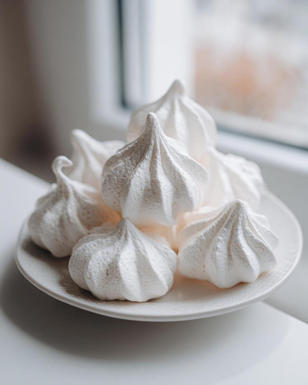 A close-up of several perfectly piped, crisp white meringue cookies stacked on a small white plate near a window.