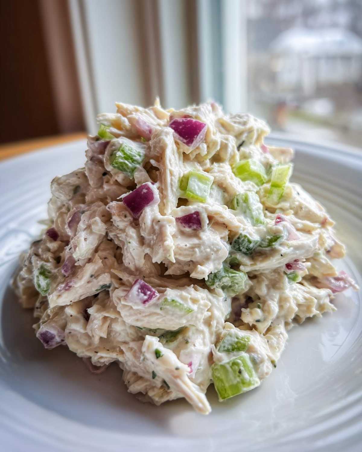 A mound of creamy greek yogurt chicken salad featuring shredded chicken, diced red onion, and celery pieces, served on a white plate.