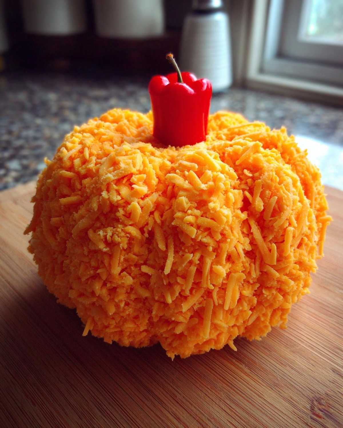 A festive pumpkin cheese ball covered in shredded cheddar cheese with a small red pepper stem.