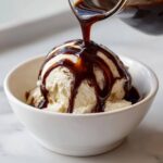 A scoop of vanilla ice cream in a white bowl being drenched with homemade chocolate syrup.