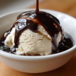 Close-up of thick, dark chocolate syrup being poured over a scoop of vanilla ice cream in a white bowl.