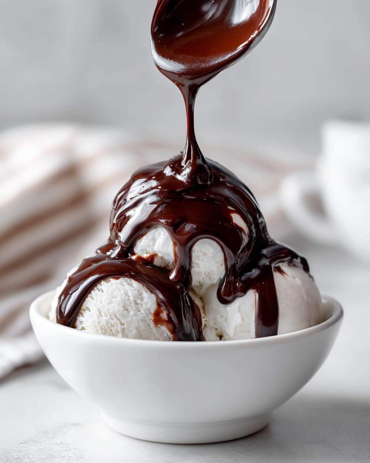 A spoonful of thick, glossy chocolate syrup is being drizzled over scoops of vanilla ice cream in a white bowl.