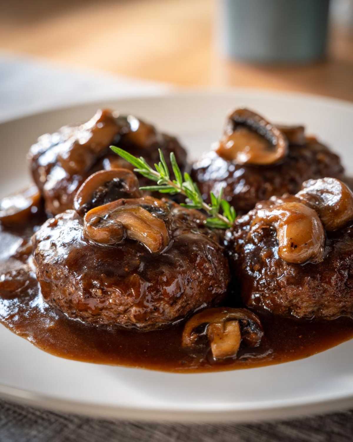 Four patties of Salisbury steak smothered in dark mushroom gravy and topped with sliced mushrooms and a rosemary sprig.