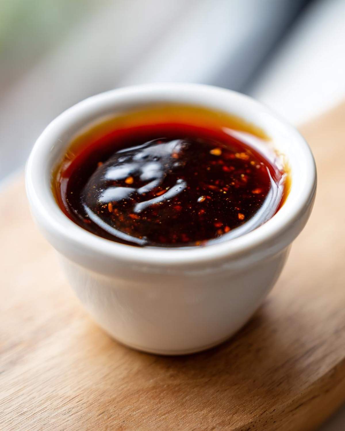 A close-up shot of thick, dark, glossy homemade teriyaki sauce speckled with chili flakes in a small white dipping bowl.