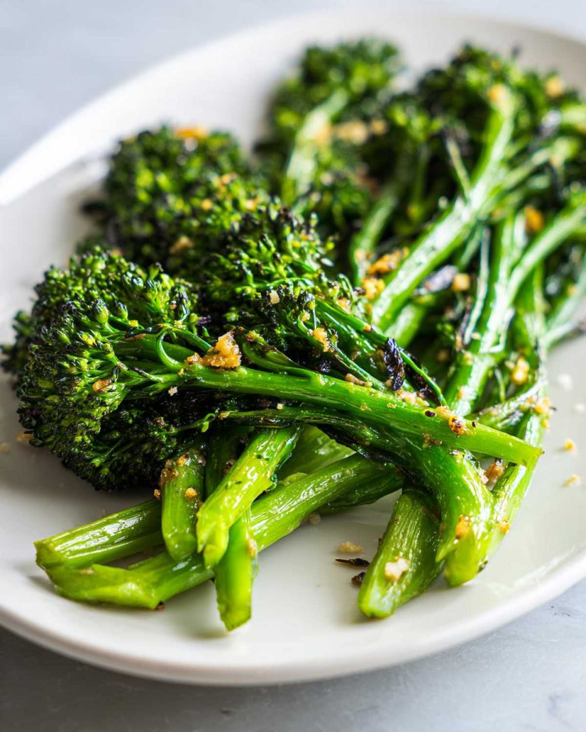 Close-up of vibrant green, roasted broccolini spears seasoned with toasted garlic bits on a white plate.