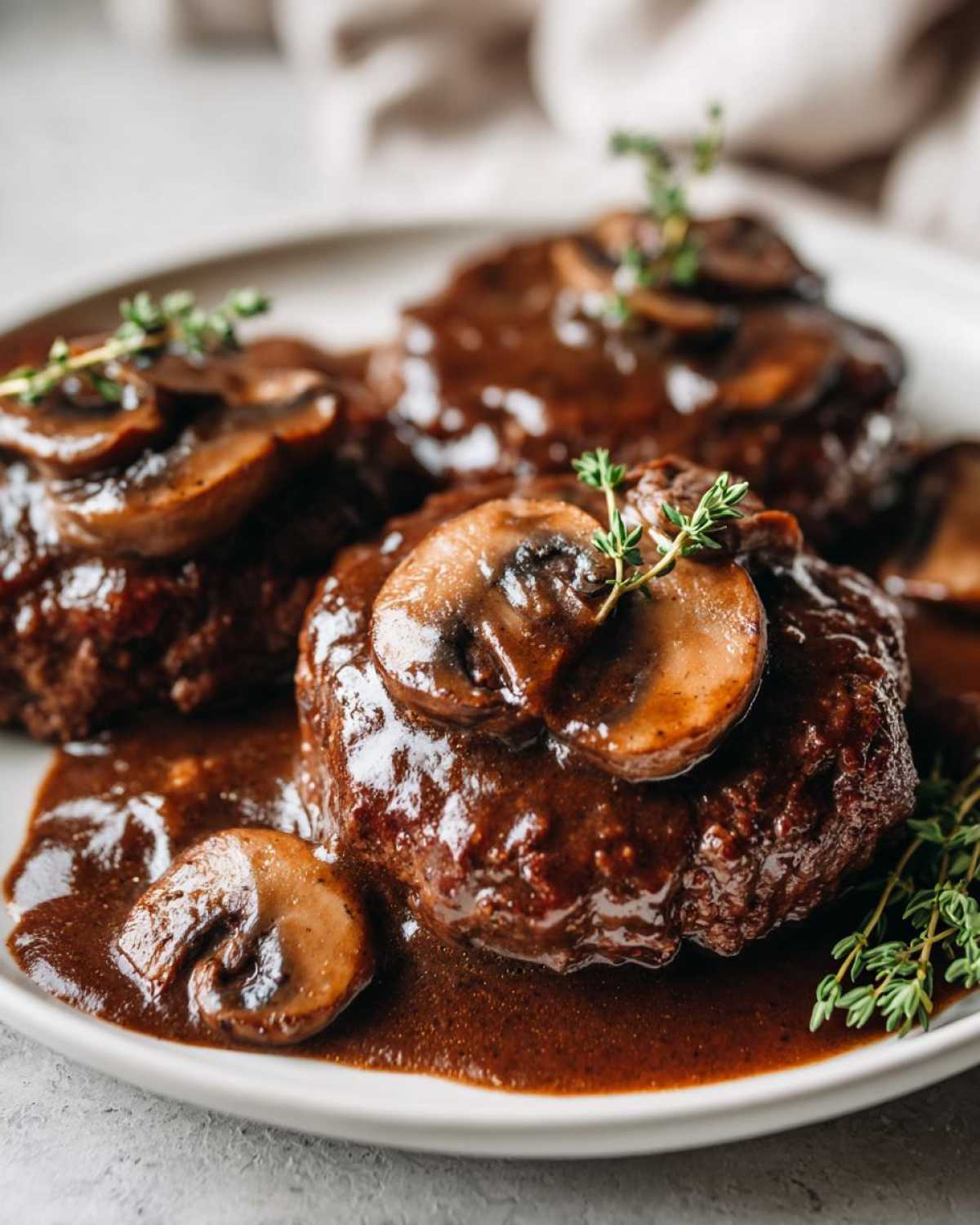 Close-up of three servings of rich Salisbury steak smothered in dark mushroom gravy and garnished with fresh thyme.
