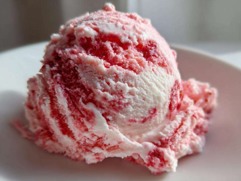 Close-up of a single scoop of homemade strawberry ice cream with visible red swirls on a white plate.