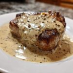 A perfectly seared steak generously topped with creamy peppercorn sauce on a white plate.