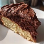 A close-up slice of vanilla cake topped with thick, rich chocolate frosting.