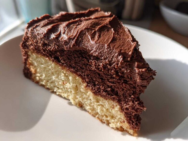 A close-up slice of vanilla cake topped with thick, rich chocolate frosting.