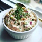 A creamy, pink smoked salmon dip garnished with fresh dill, red onion, and capers in a white ramekin.