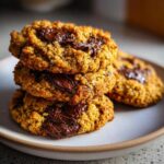 A stack of three golden brown sourdough chocolate chip cookies piled high on a light plate, featuring melted chocolate.