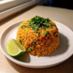 A mound of vibrant orange Spanish rice mixed with green peas, topped with fresh cilantro, served with a lime wedge.
