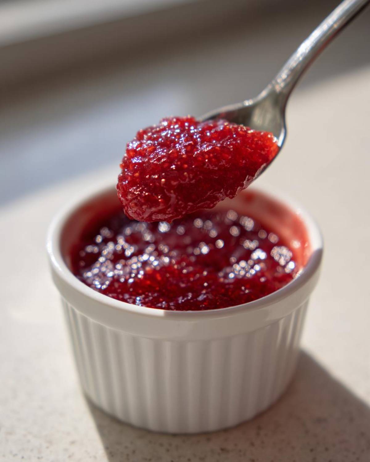 A spoonful of glistening, bright red homemade raspberry jam being lifted from a small white ramekin.