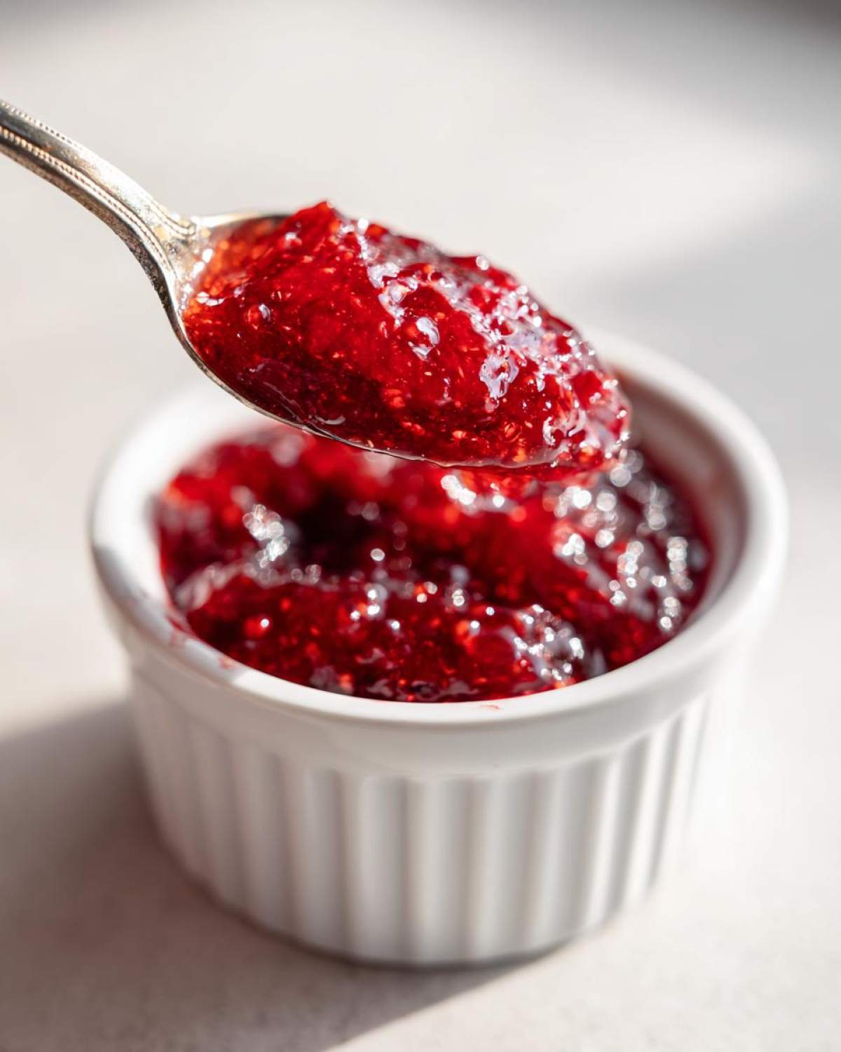 A silver spoon lifting a spoonful of glistening, bright red raspberry jam above a small white ramekin.