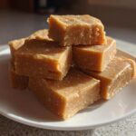 A stack of rich, square pieces of homemade penuche fudge sitting on a white plate bathed in sunlight.