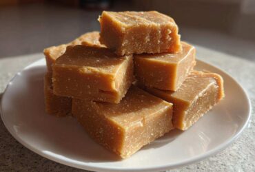 A stack of rich, square pieces of homemade penuche fudge sitting on a white plate bathed in sunlight.