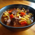 Close-up of a steaming bowl of rich beef noodle soup featuring chunks of beef, egg noodles, carrots, and celery.
