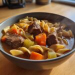 Close-up of a steaming bowl of rich beef noodle soup featuring tender beef chunks, egg noodles, carrots, and celery.