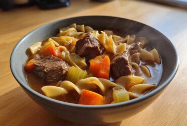 Close-up of a steaming bowl of rich beef noodle soup featuring tender beef chunks, egg noodles, carrots, and celery.
