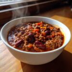 Close-up of a steaming white bowl filled with rich, hearty chili featuring ground beef, kidney beans, and diced vegetables.