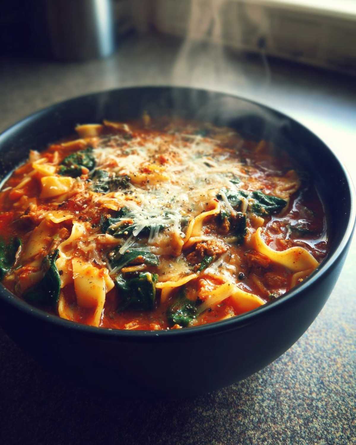 Close-up of a steaming black bowl filled with cheesy one pot lasagna soup, featuring noodles, spinach, and melted cheese.