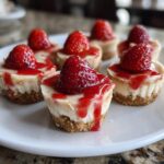 A close-up of several delicious strawberry cheesecake bites topped with fresh strawberries and glaze on a white plate.
