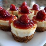 A close-up of several individual strawberry topped cheesecake bites arranged on a white serving platter.