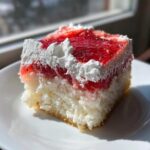 A close-up slice of moist jello poke cake featuring white cake, bright red strawberry jello, and swirls of whipped cream topping.