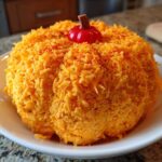 A vibrant orange pumpkin cheese ball coated entirely in shredded cheddar cheese and topped with a small decorative stem.