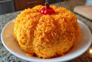 A vibrant orange pumpkin cheese ball coated entirely in shredded cheddar cheese and topped with a small decorative stem.