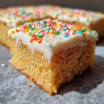 A close-up of a square serving of soft sugar cookie bars topped with thick white frosting and colorful rainbow sprinkles.