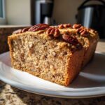 Close-up of a moist slice of sweet Alabama pecan bread topped with whole pecans, sitting on a white plate.